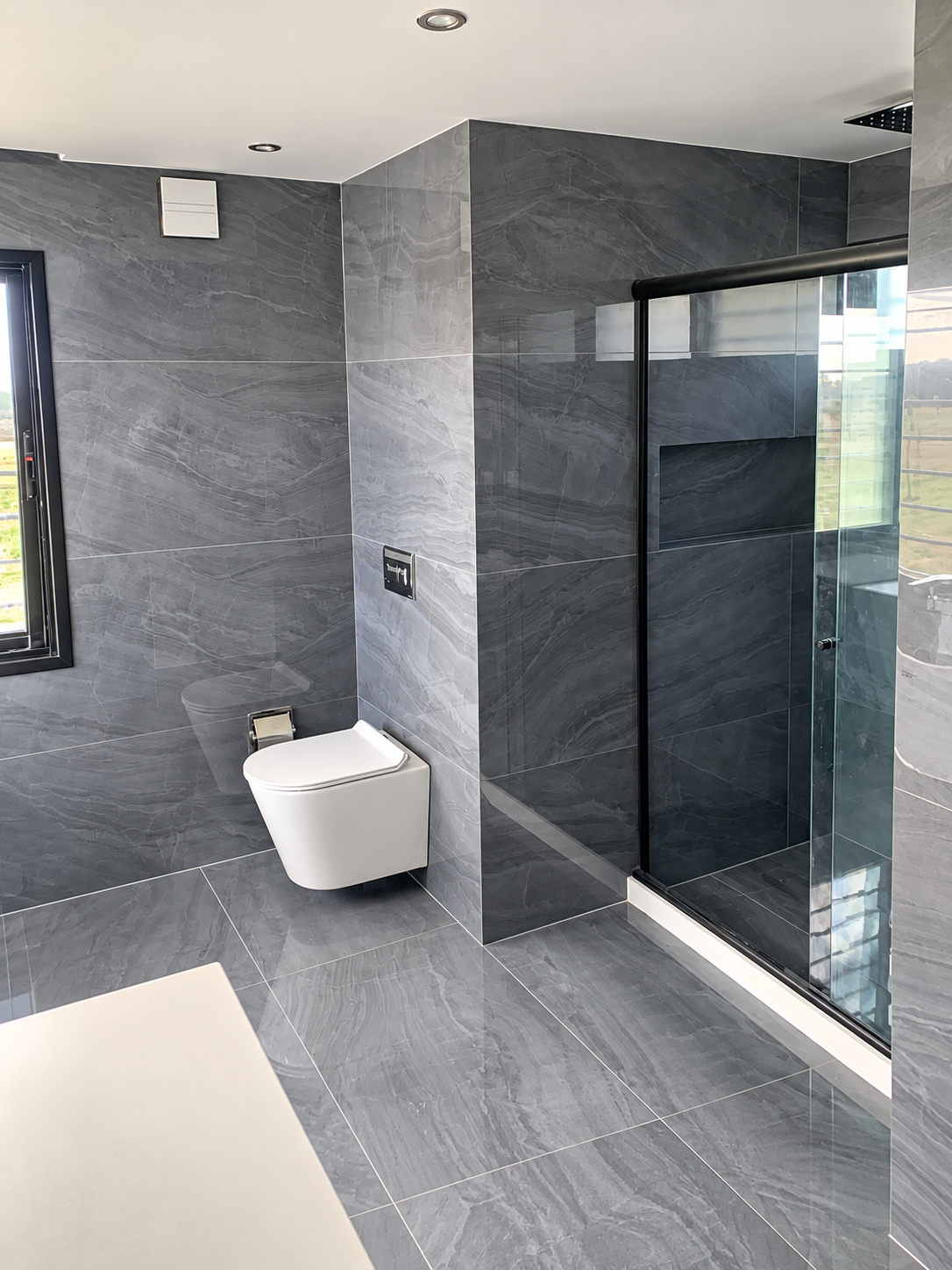 Baño reformado con revestimiento gris, mampara de vidrio y perfiles negros.